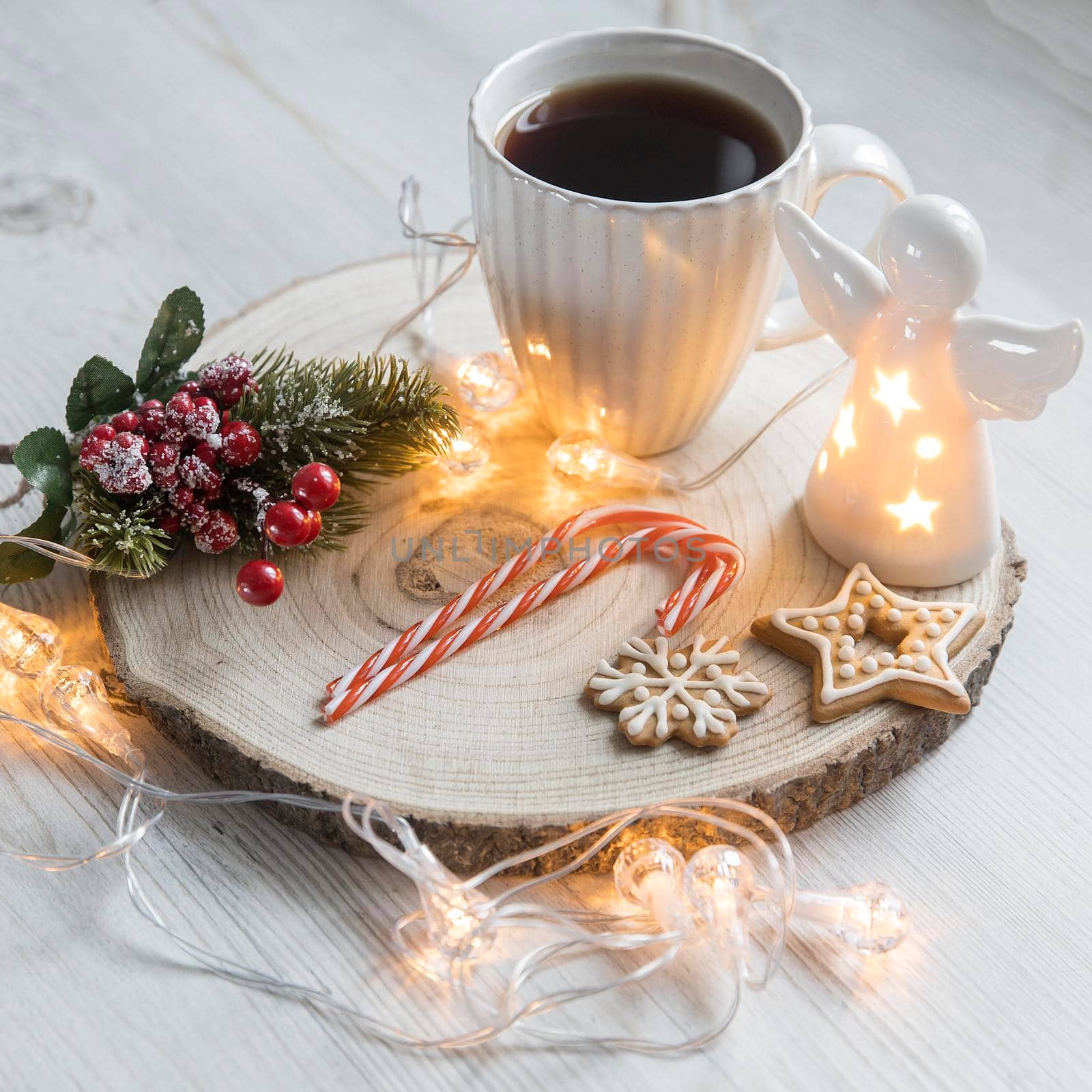 A cup of coffee on a wooden stand, a gingerbread cookie tied with a rope, a garland on the table. Artificial spruce branch with red berries. Breakfast at Christmas. Scandinavian style. Copy space by elenarostunova