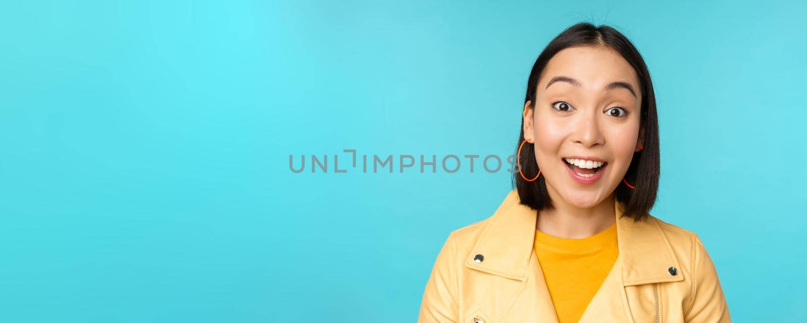 Close up portrait of natural asian girl laughing, smiling and looking happy, standing over blue background. Copy space