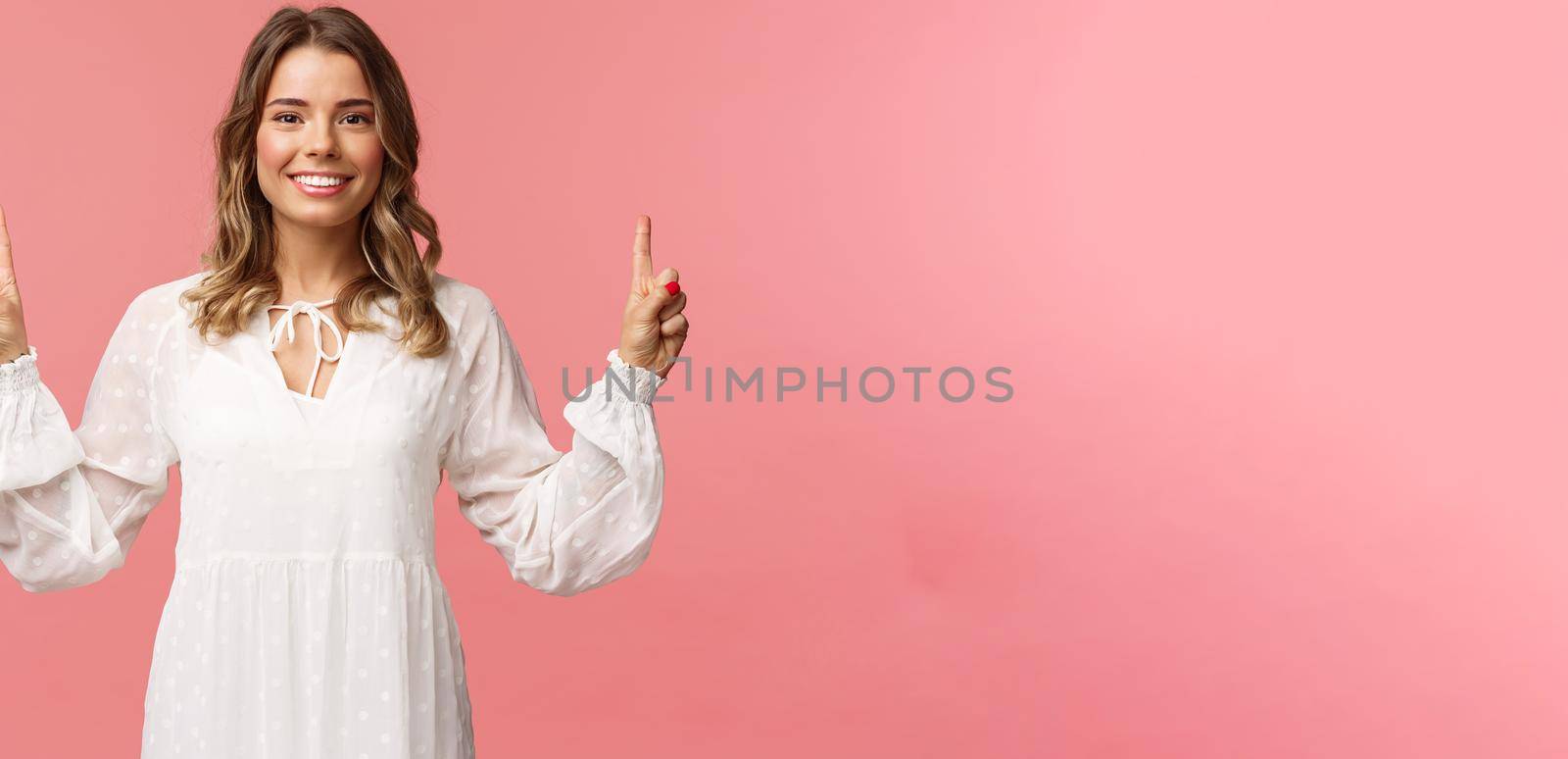 Portrait of confident beautiful young blond woman in white cute dress, pointing fingers up at top advertisement, looking at camera with beaming smile, standing pink background.