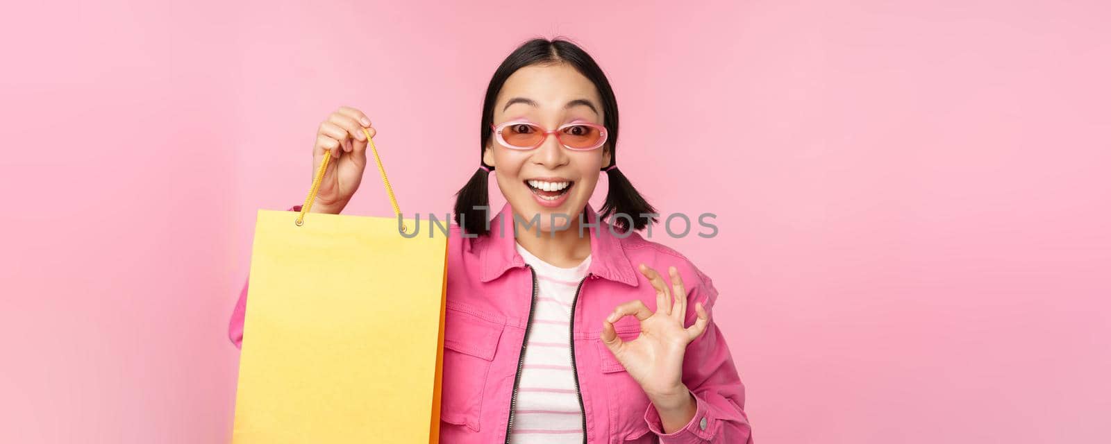 Shopping. Stylish asian girl in sunglasses, showing bag from shop and smiling, recommending sale promo in store, standing over pink background.
