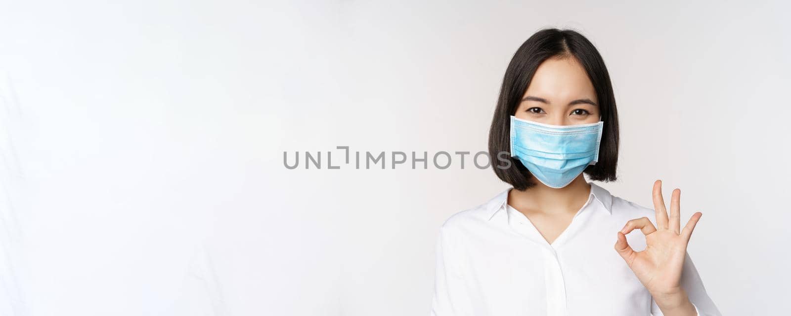 Covid and health concept. Portrait of asian woman wearing medical face mask and showing okay sign, standing over white background.