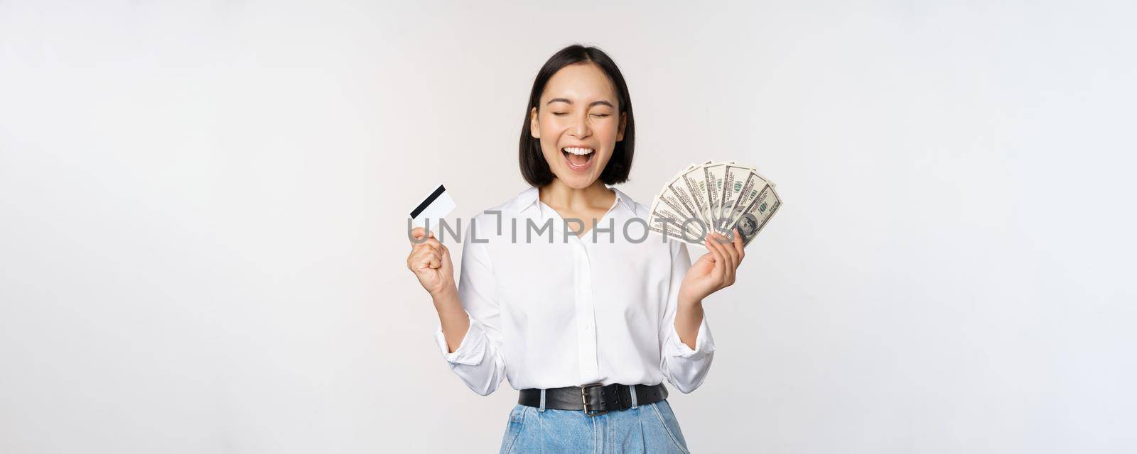 Finance and money concept. Happy young asian woman dancing with cash and credit card, smiling pleased, posing against white studio background.