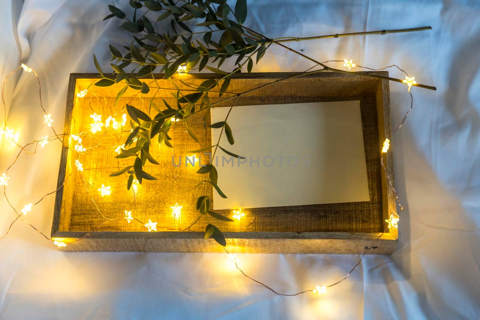 A eucalyptus branch lies on a white sheet beside a tray with a cup of tea and a saucer in a letter in an envelope. The garland is glowing. by elenarostunova