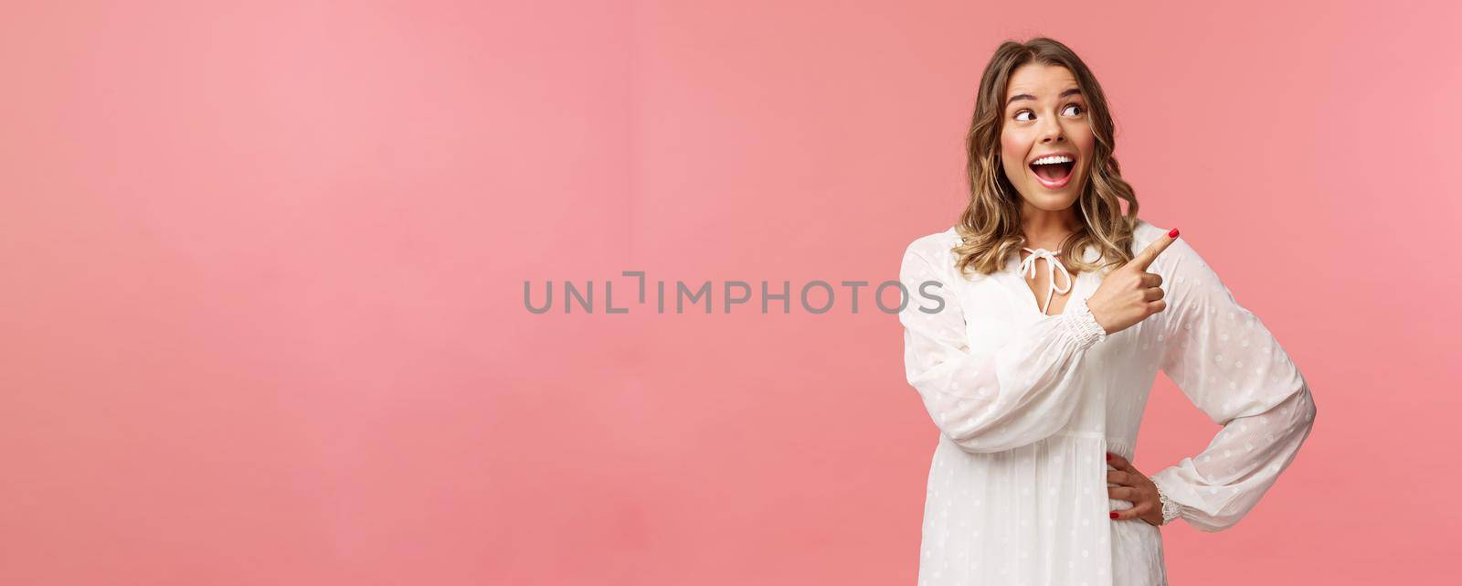 Enthusiastic, happy attractive blond woman in white cute dress, open mouth excited and cheerful looking, pointing upper right corner as best prices discounts ever, stand pink background.