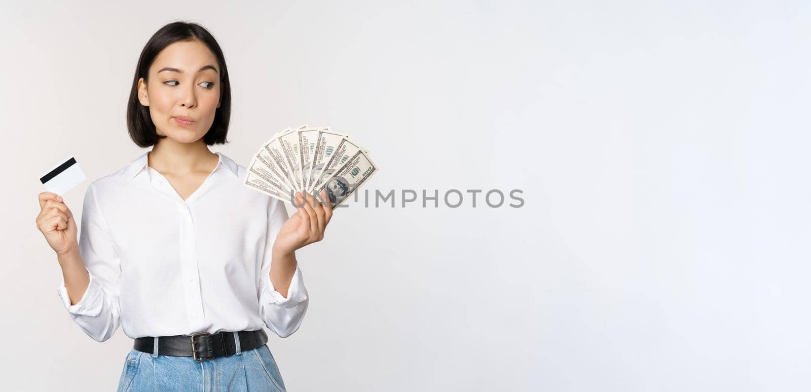 Image of asian woman looking at money dollars, holding credit card in another hand, thinking, standing over white background.