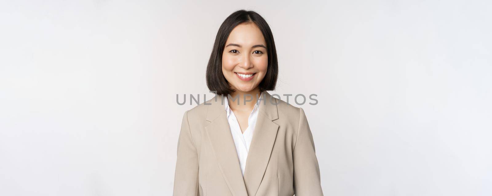 Portrait of successful businesswoman in suit, smiling and looking like professional at camera, white background by Benzoix