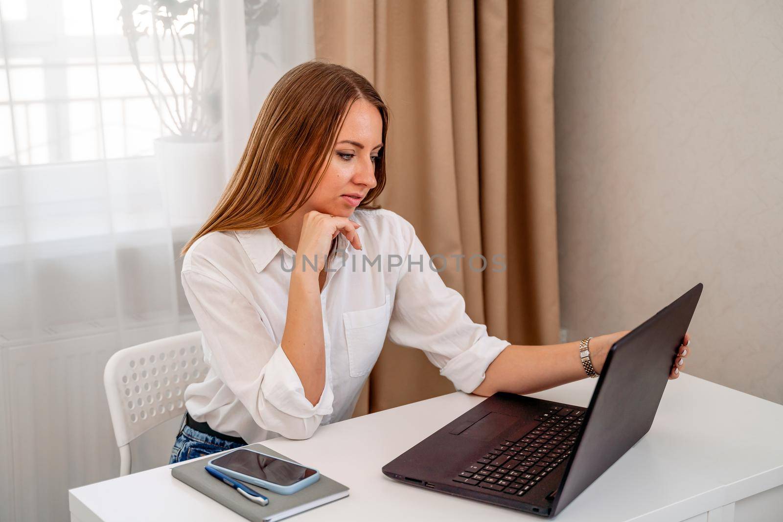 European professional woman is sitting with a laptop at a table in a home office, a positive woman is studying while working on a PC. She is wearing a beige jacket and jeans and is on the phone. by Matiunina