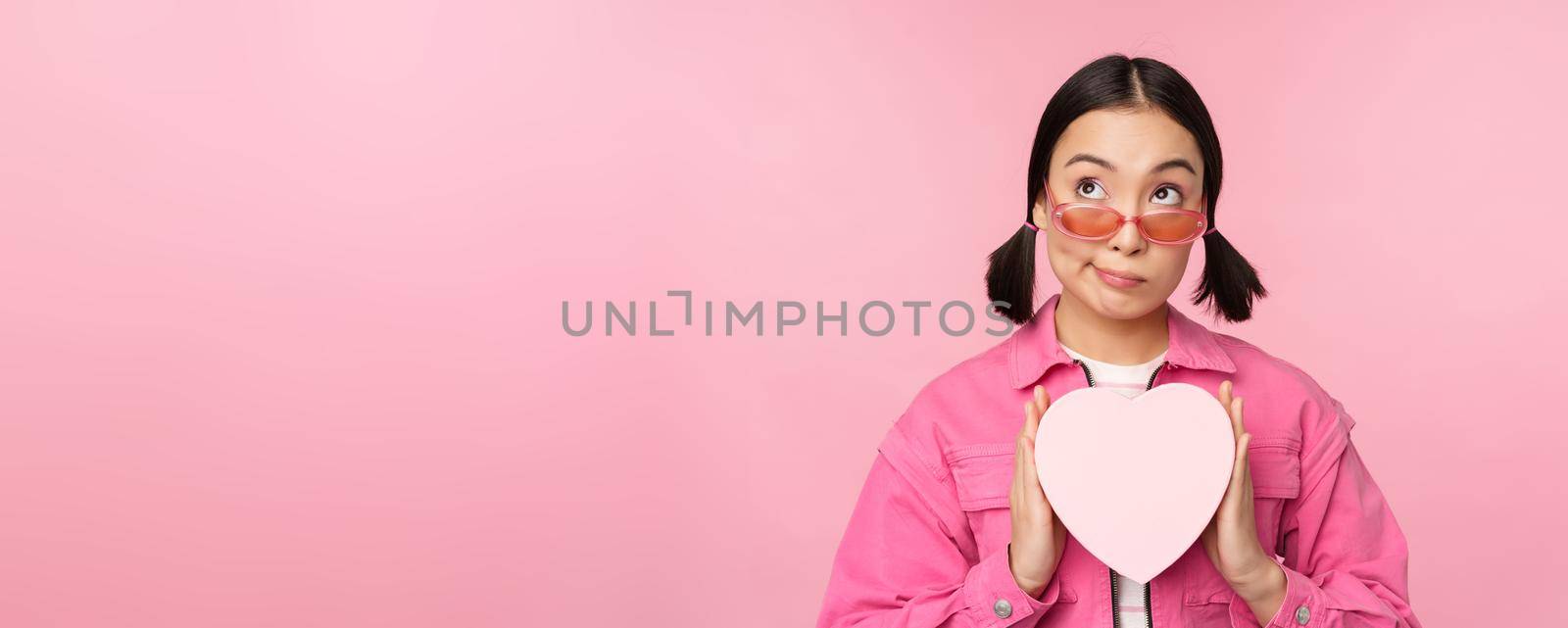 Beautiful asian girl smiling happy, showing heart gift box and looking excited at camera, standing over pink romantic background.