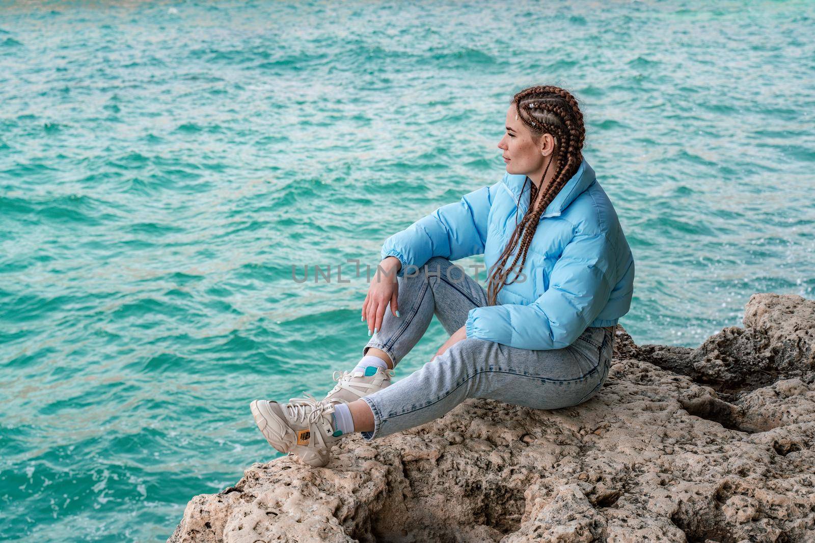 A woman in a blue jacket sits on a rock above a cliff above the sea, looking at the stormy ocean. Girl traveler rests, thinks, dreams, enjoys nature. Peace and calm landscape, windy weather