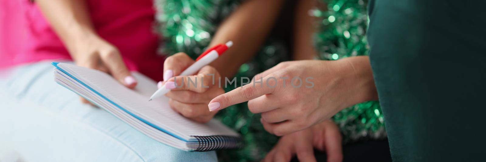 Close-up of women writing wish list for christmas, empty page to list desires for gifts. Creative atmosphere before new year, presents under christmas tree. Holiday concept. Blurred background