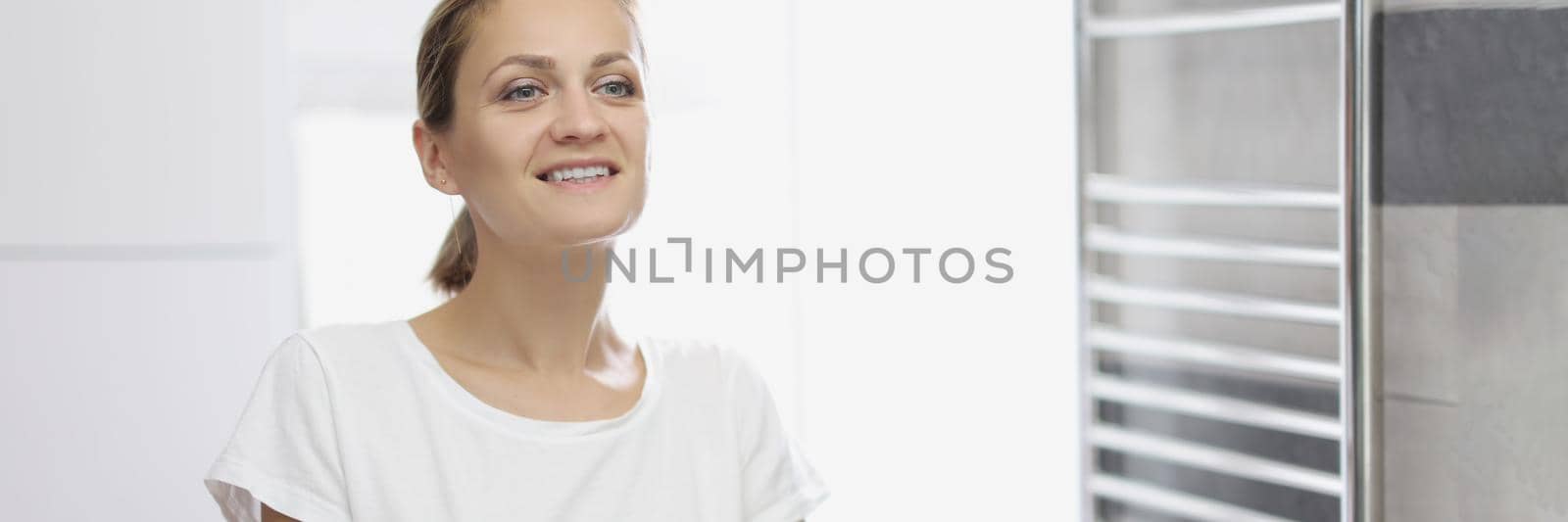 Portrait of young woman looking in mirror at herself in bathroom, white smile, natural beauty, no makeup, morning routine, light restroom. Good morning, wellness, beauty, youth concept