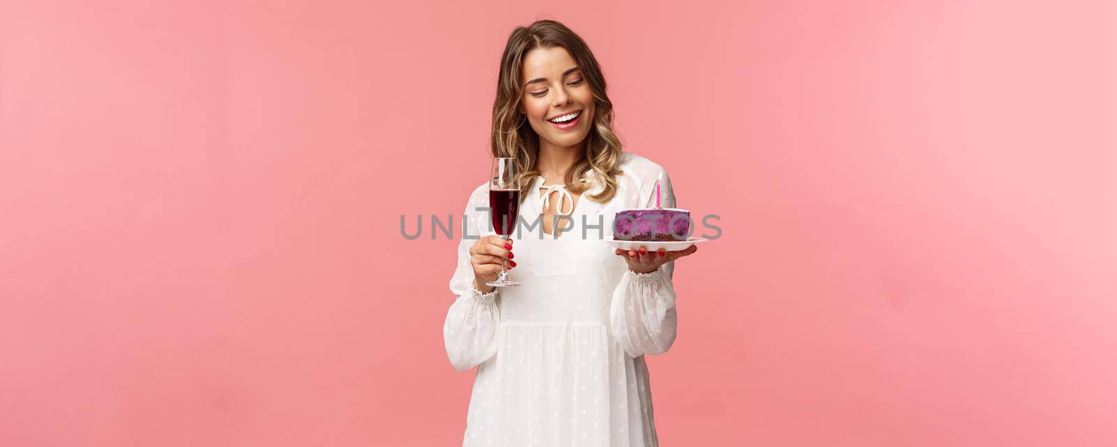 Holidays, spring and party concept. Portrait of tender, elegant young blond woman holding glass of wine and cake with lit candle, smiling pleased, celebrating birthday, pink background.