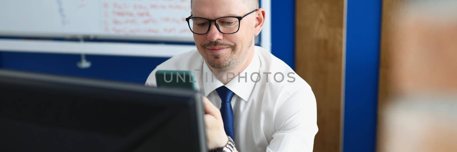 Portrait of company boss texting in smartphone, leader in private office. Computer on workplace, board with lettering on wall. Business, working day, career growth, technology concept