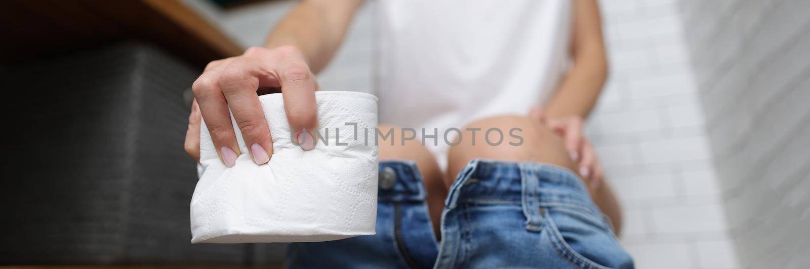 Close-up of woman taking toilet paper to use for hygiene. Bog roll for cleaning body, female on toilet seat. Bathroom, restroom, nature calls concept. Blurred background