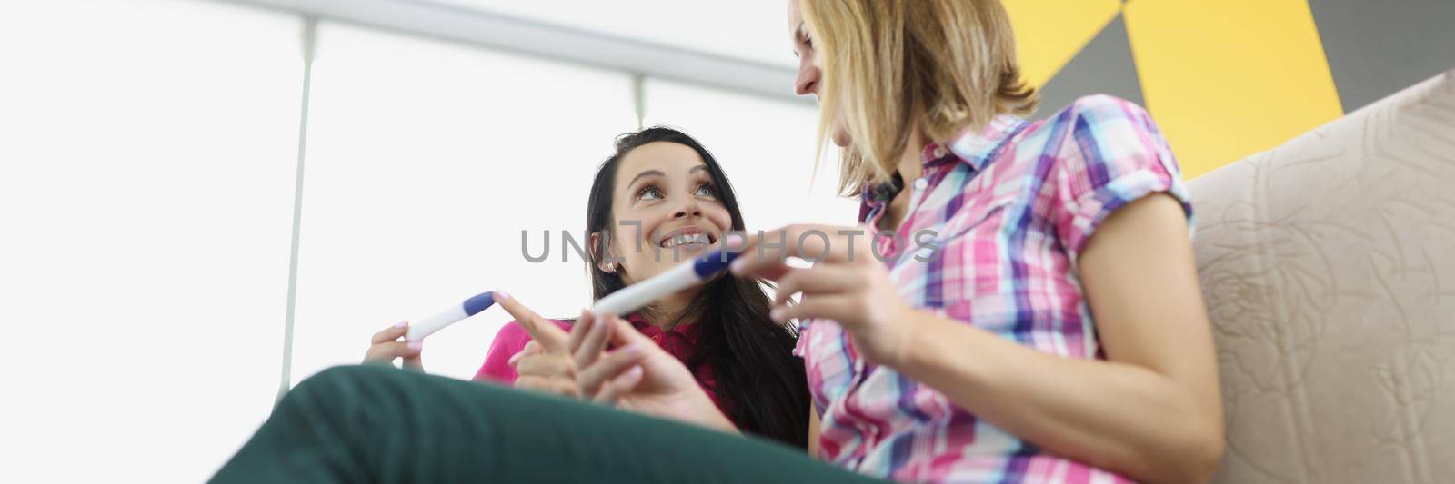 Low angle of friends girls hold pregnancy tests, happy best friends got big news. Smiling women on sofa at home. Friendship, pregnancy, new period, life change, happiness concept