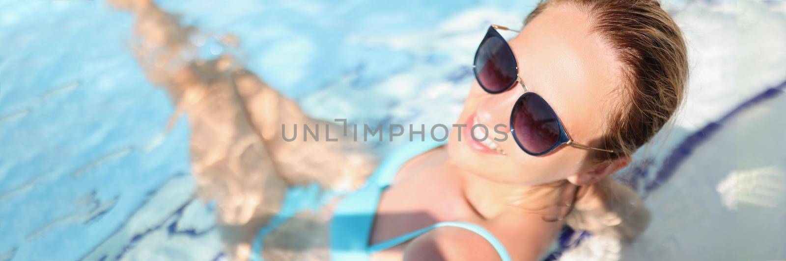 Portrait of carefree young woman relaxing in luxury swimming pool, smiling female wear glasses, blue swimsuit, clean water. Holiday, summer, resort, chilling, vacation concept