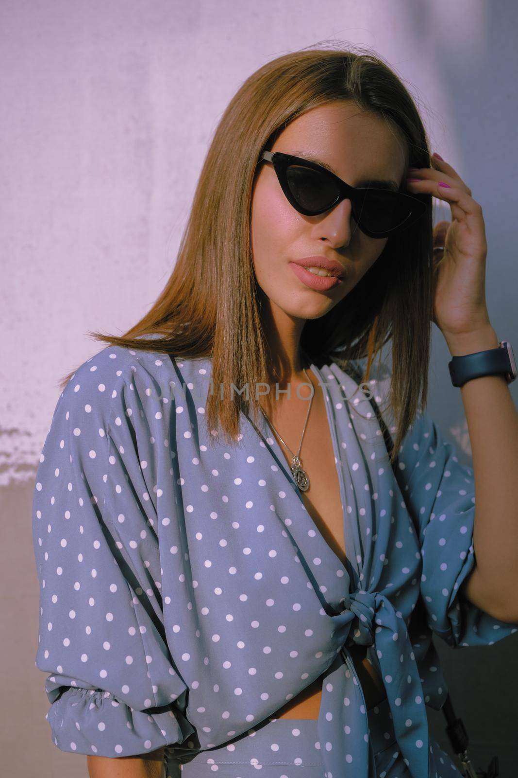 Gorgeous blonde maiden in a long blue dress with polka-dots, watch, sunglasses, with a pendant around her neck and a small black handbag on her shoulder is touching her hair and looking at the camera while walking alone in the city. The concept of fashion and style. Close-up shot.