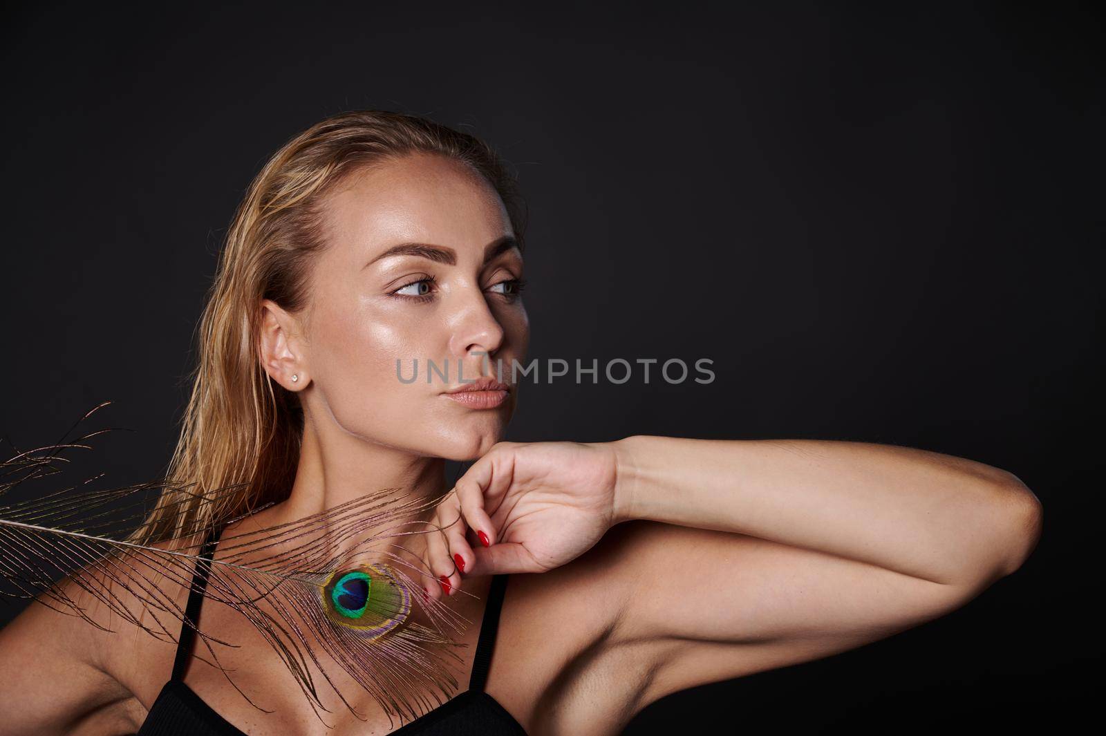 Close-up gorgeous stunning middle aged blonde Caucasian woman with healthy fresh clean skin and wet blond hair holding a peacock feather, isolated over black background with copy ad space