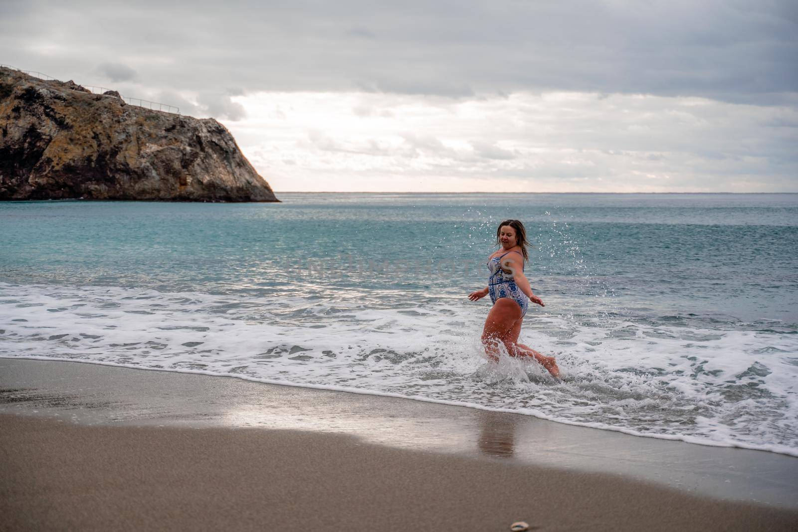 A plump woman in a bathing suit enters the water during the surf. Alone on the beach, Gray sky in the clouds, swimming in winter