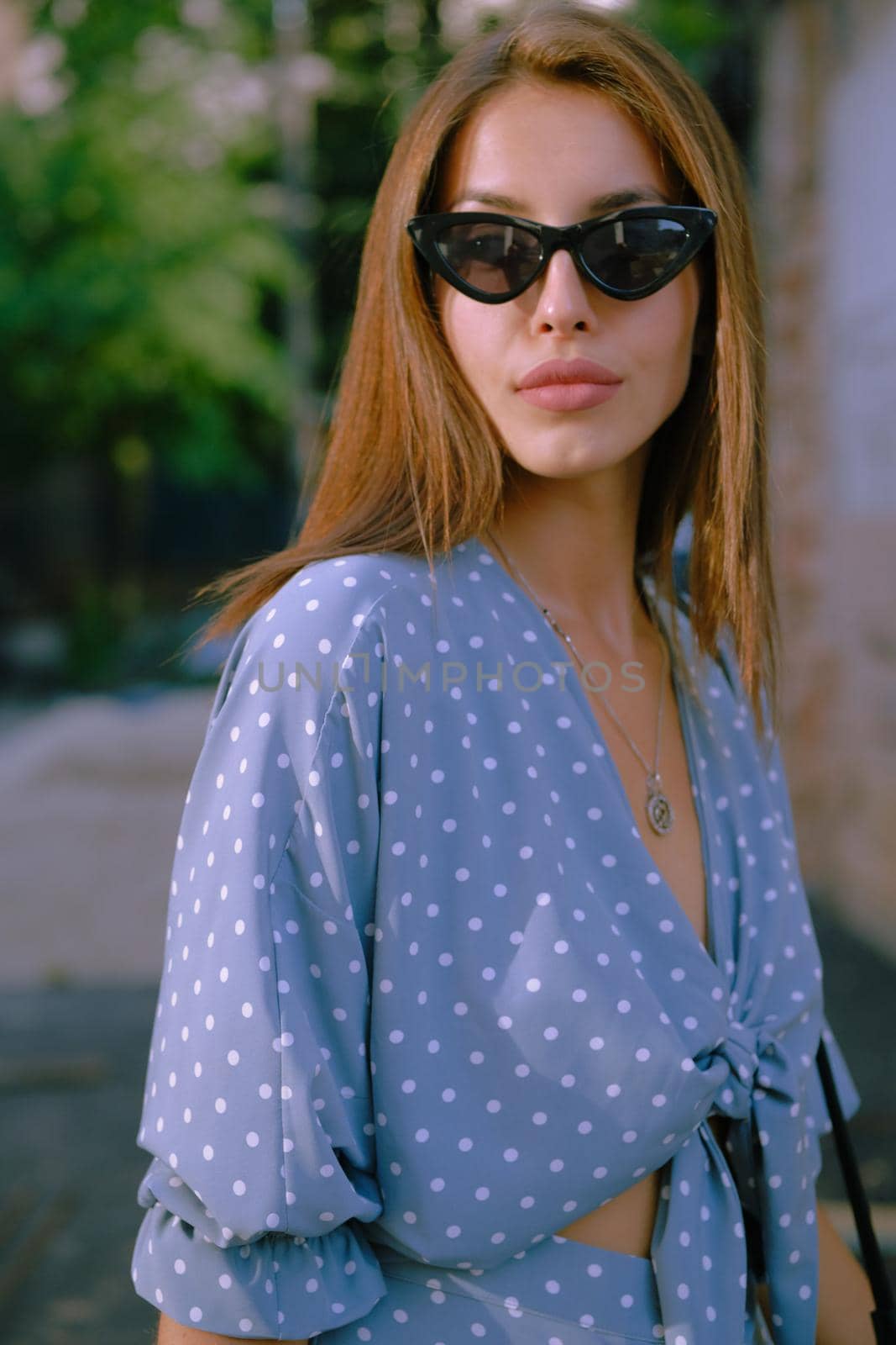 Charming blonde woman in a long blue dress with polka-dots, watch, sunglasses, with a pendant around her neck and a small black handbag on her shoulder is walking alone in the city. The concept of fashion and style. Close-up shot.
