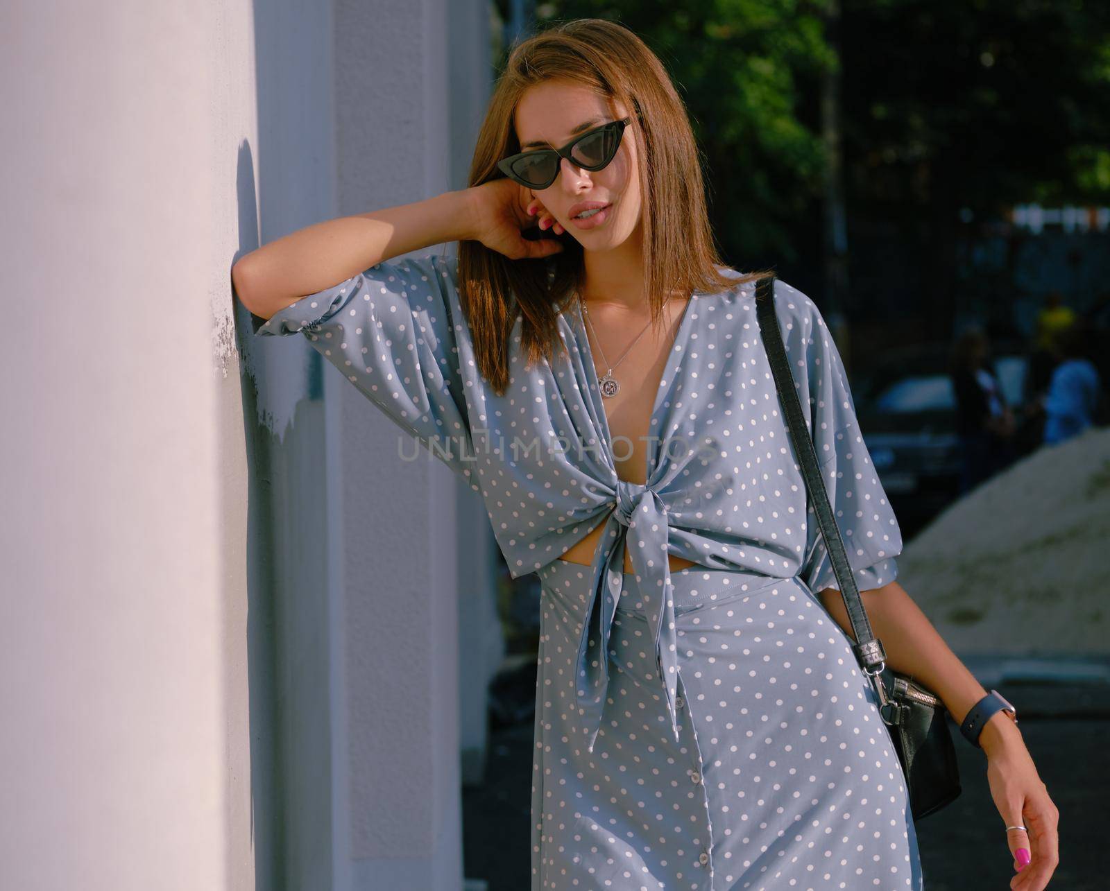 Gorgeous blonde lady in a long blue dress with polka-dots, watch, sunglasses, with a pendant around her neck and a small black handbag on her shoulder is posing leaning her elbow on a wall and looking at the camera while walking alone in the city. The concept of fashion and style. Close-up shot.