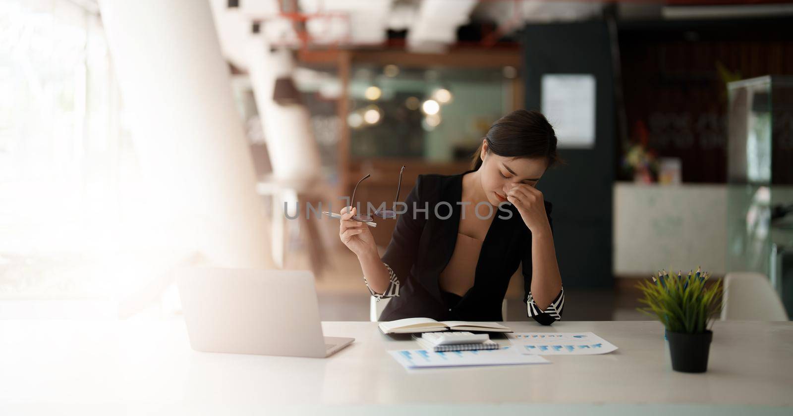 Very stressed business asian woman sitting in front of her computer looking at a large pile of paperwork, while holding a hand at her forehead.