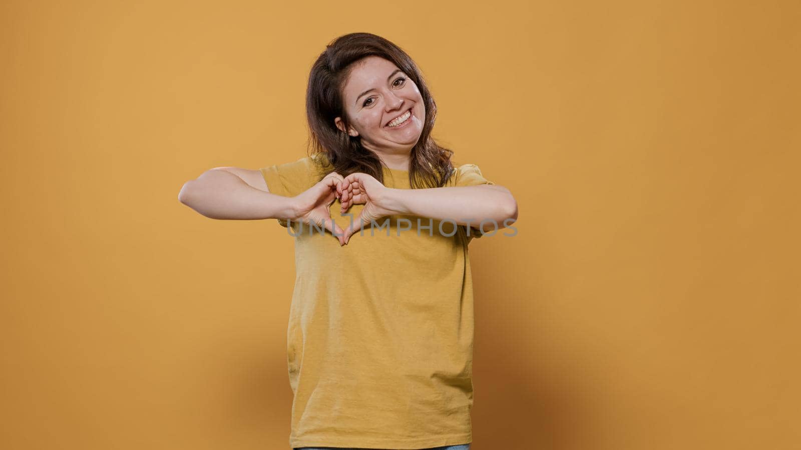 Happy woman gesturing making heart sign with hands to show loving feelings for her partner over yellow background. Smiling person feeling in love talking romantic for valentine day.