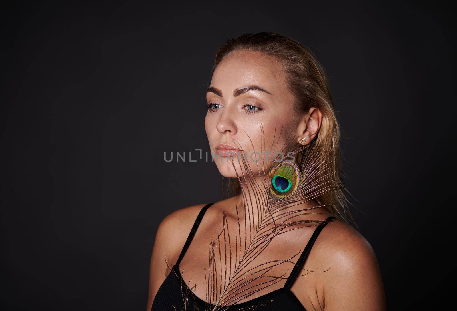 Attractive middle aged blonde woman with healthy fresh clean tanned radiant skin holding a peacock feather, isolated over black background with copy ad space