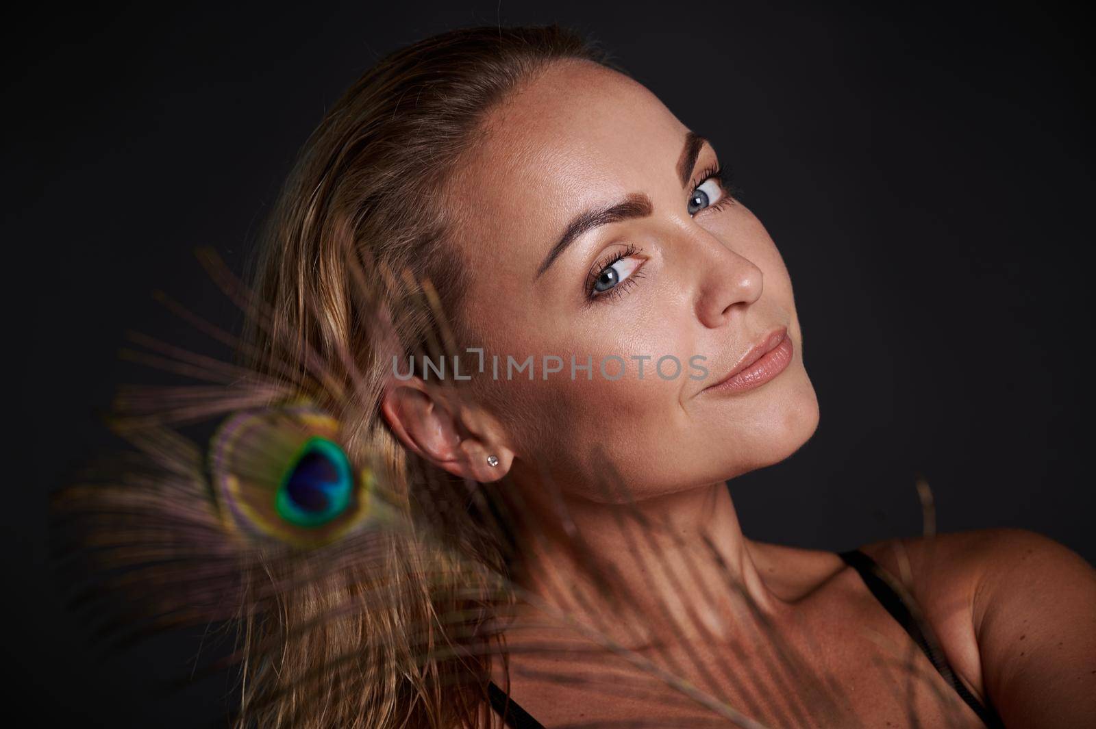 Headshot gorgeous smiling stunning middle aged blonde Caucasian woman with healthy fresh clean skin and wet blond hair holding a peacock feather, isolated over black background with copy ad space by artgf