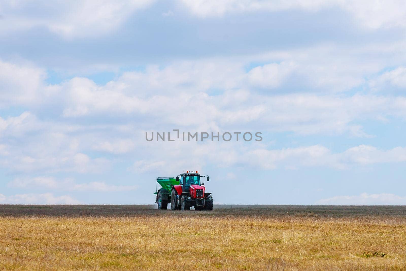 A red tractor in a green trailer is carrying fertilizer to the field. Agricultural work is performed by a farmer.