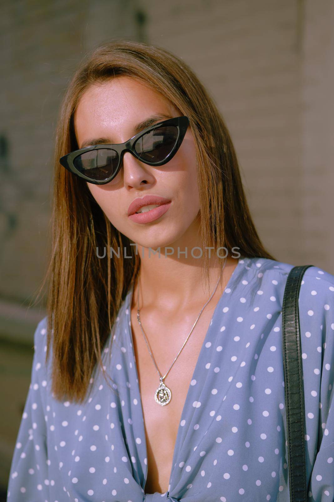 Beautiful blonde lady in a long blue dress with polka-dots, watch, sunglasses, with a pendant around her neck and a small black handbag on her shoulder is looking at the camera while walking alone in the city. The concept of fashion and style. Close-up shot.