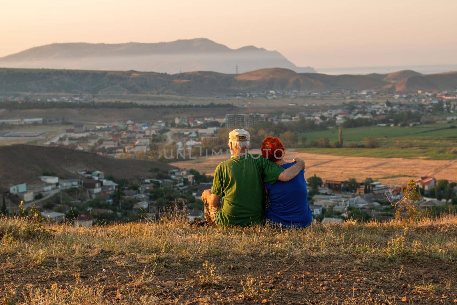 An elderly couple sits on a mountain with their backs with a beautiful view of the mountains and the sea in the distance