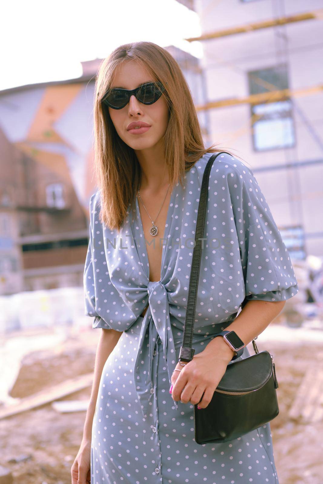 Pretty blonde girl in a long blue dress with polka-dots, watch, sunglasses, with a pendant around her neck and a small black handbag on her shoulder is looking at the camera while walking alone in the city. The concept of fashion and style. Close-up shot.