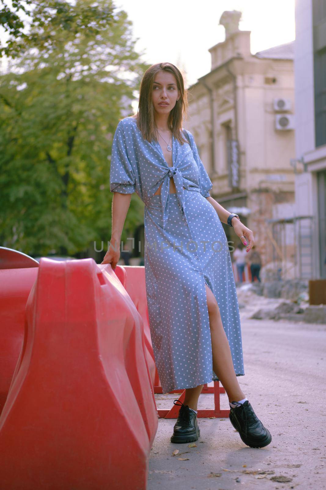 Cute blonde female in a long blue dress with polka-dots, black boots, watch, with a pendant around her neck and a small handbag on her shoulder is posing leaning a red guardrail while walking alone in the city. The concept of fashion and style. Full length shot.