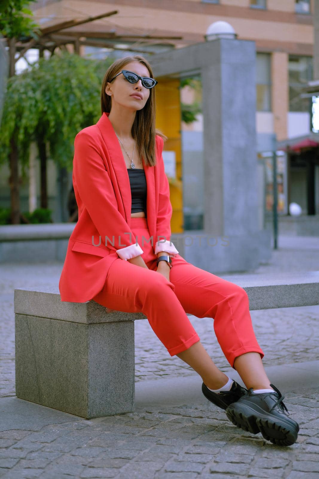 Pretty blonde female in a red lady-type pantsuit and black top, boots, watch, ring, sunglasses, with a pendant around her neck is looking up, posing sitting sideways on a stone bench while walking alone in the city. The concept of fashion and style. Full-length shot.