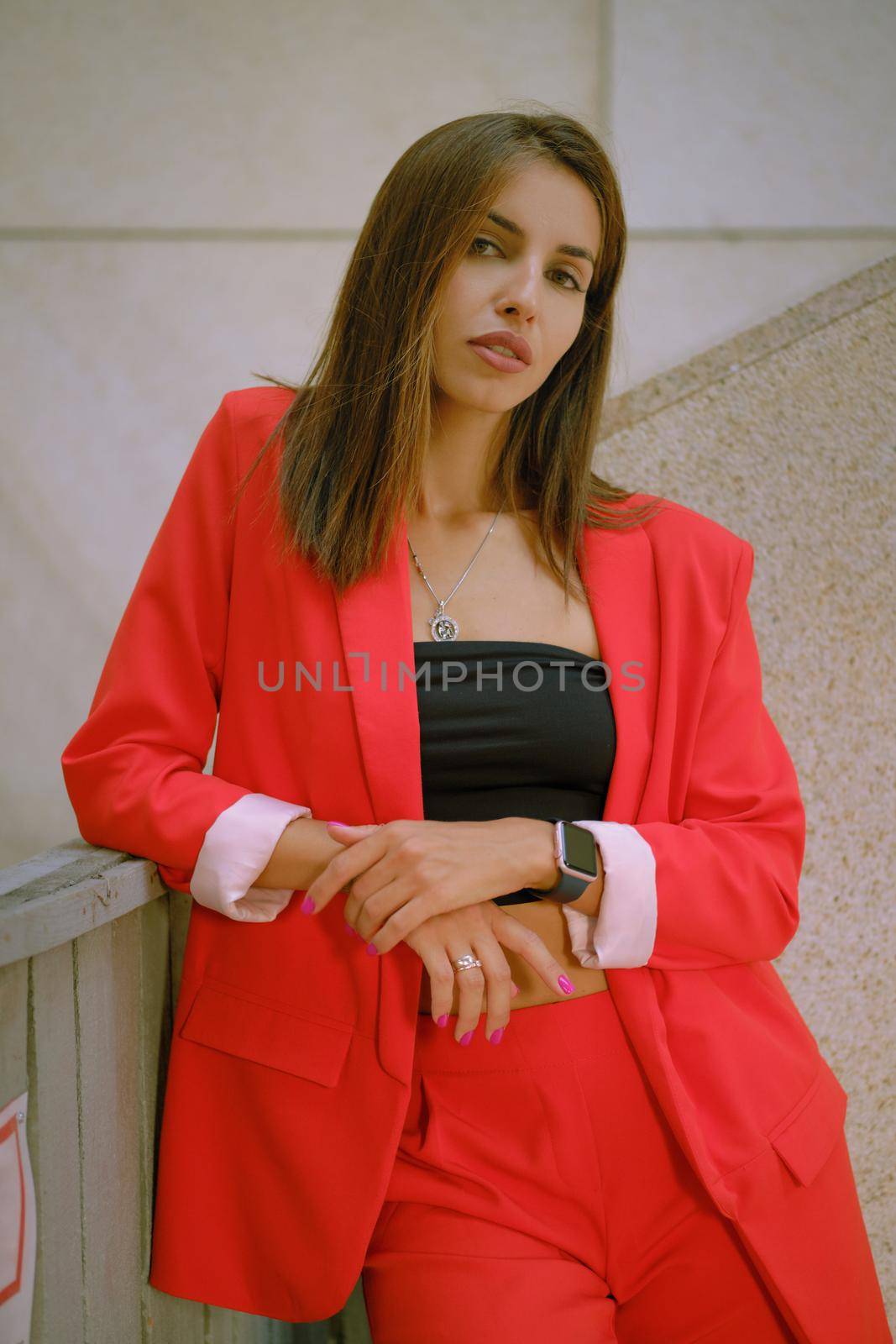 Charming blonde girl in a red lady-type pantsuit and black top, watch, ring, with a pendant around her neck is looking at the camera while walking alone in the city. The concept of fashion and style. Close-up shot.