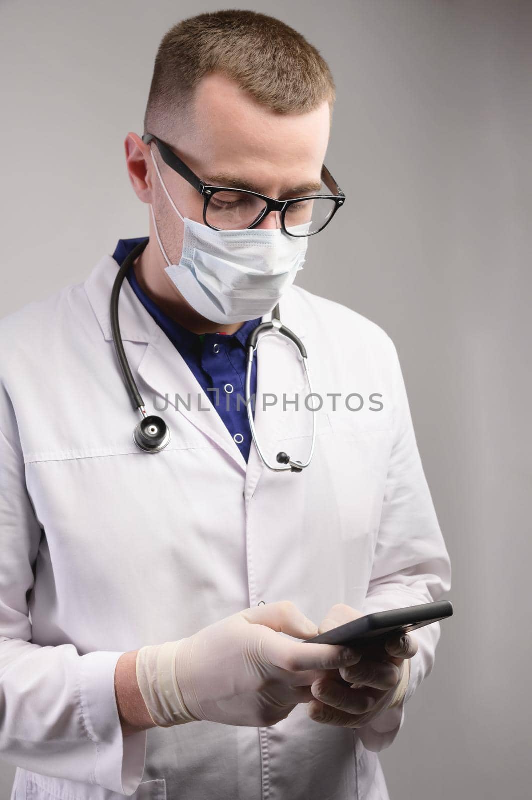 Studio portrait of a young caucasian doctor in uniform and a mask with an smartphone in his hands on a white background with copy space in the frame. by yanik88
