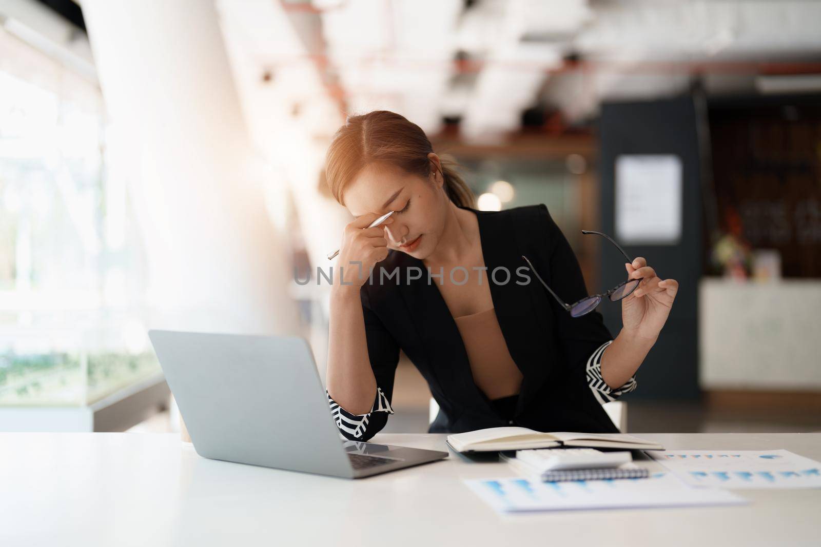 Very stressed business asian woman sitting in front of her computer looking at a large pile of paperwork, while holding a hand at her forehead.