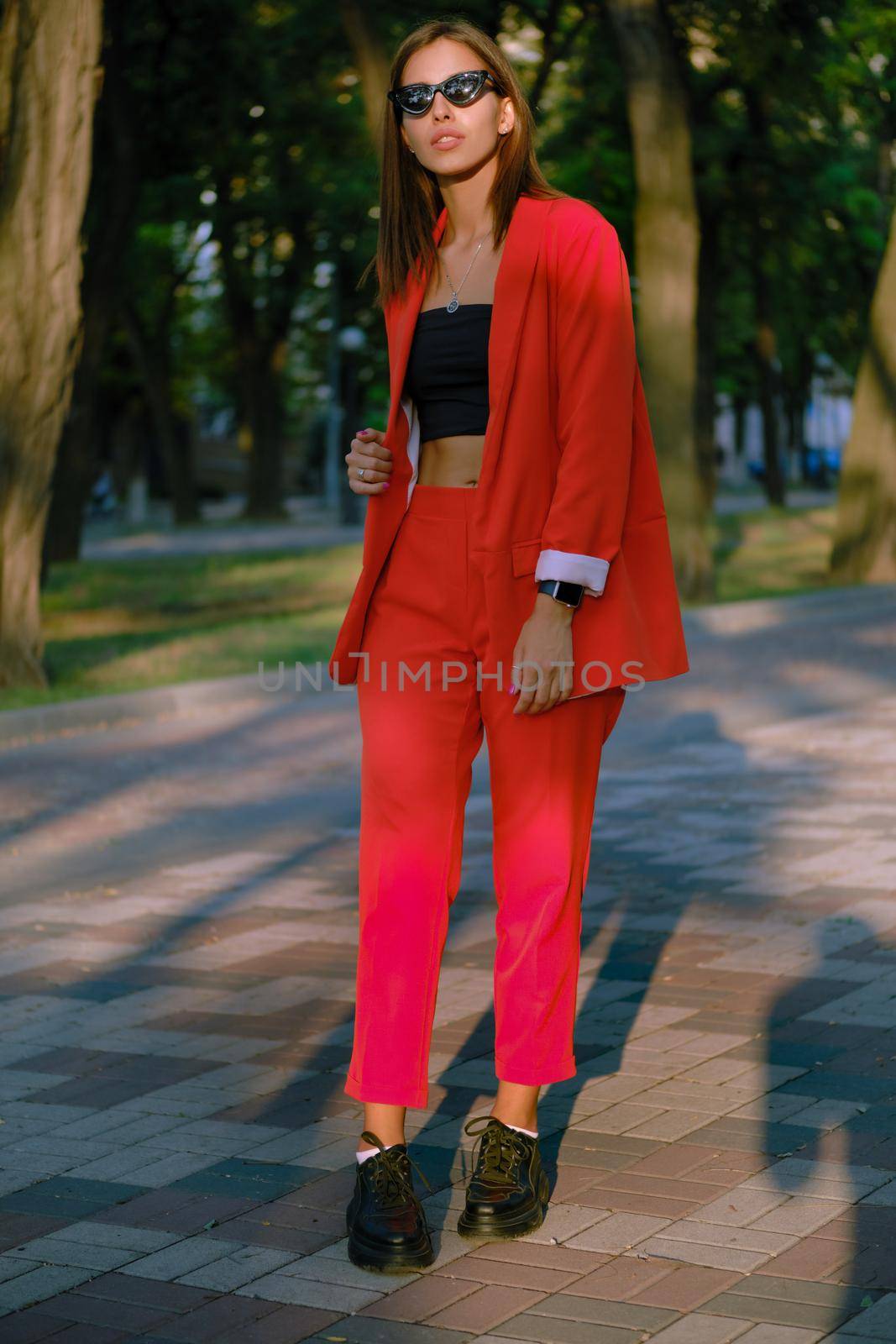 Wonderful blonde maiden in a red lady-type pantsuit and black top, boots, watch, ring, sunglasses, with a pendant around her neck is walking alone in the city. The concept of fashion and style. Full-length shot.