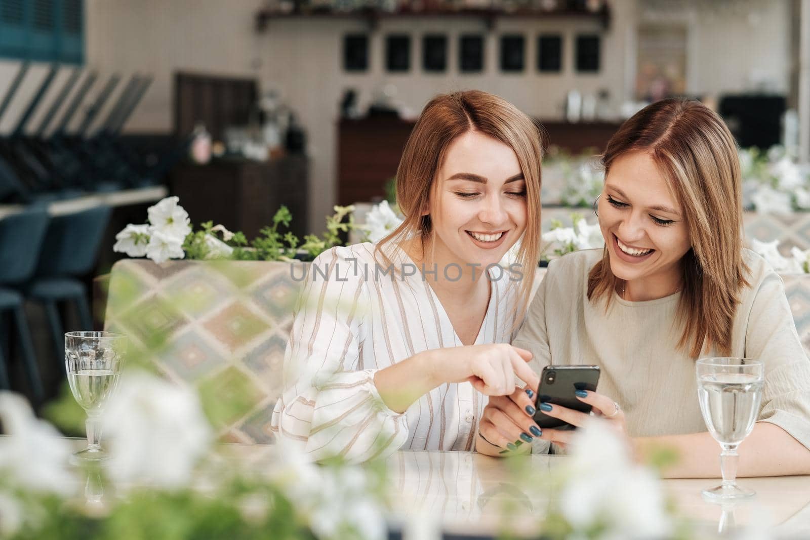Two Cheerful Women Having Lunch Meeting in the Restaurant, Watching on Smartphone and Smiling by Romvy