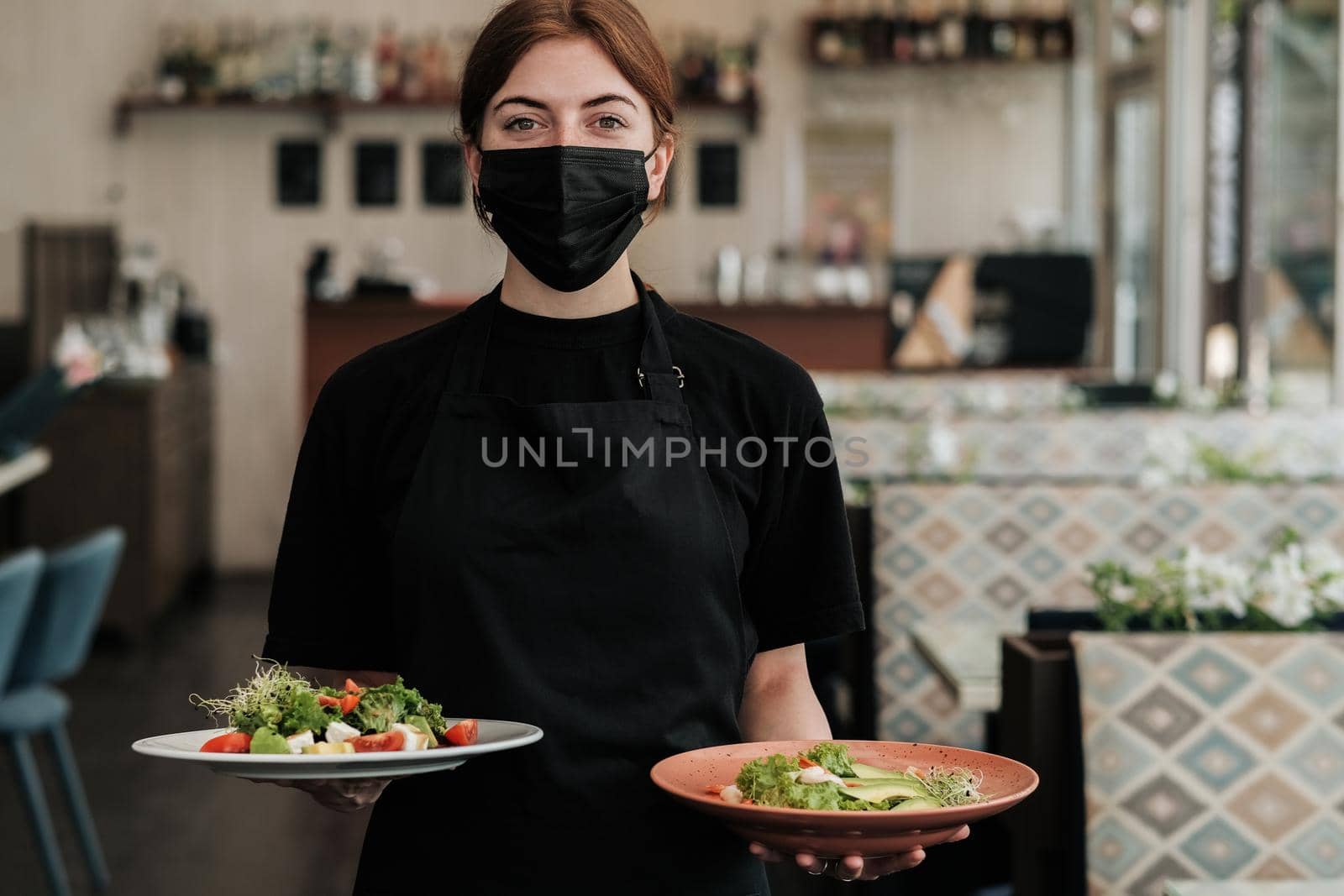 Portrait of Young Female Waiter Wearing Protection Mask that Holding Two Plates of Fresh Salad in the Restaurant by Romvy