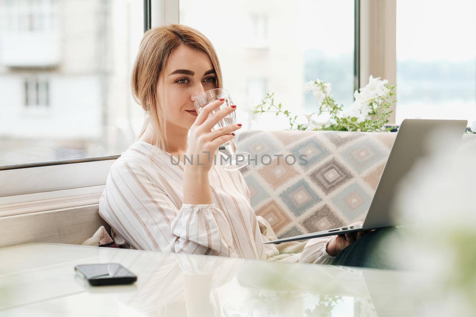 Young Woman Holding Laptop and Drinking Water While Sitting in Restaurant,Female Freelancer at Work