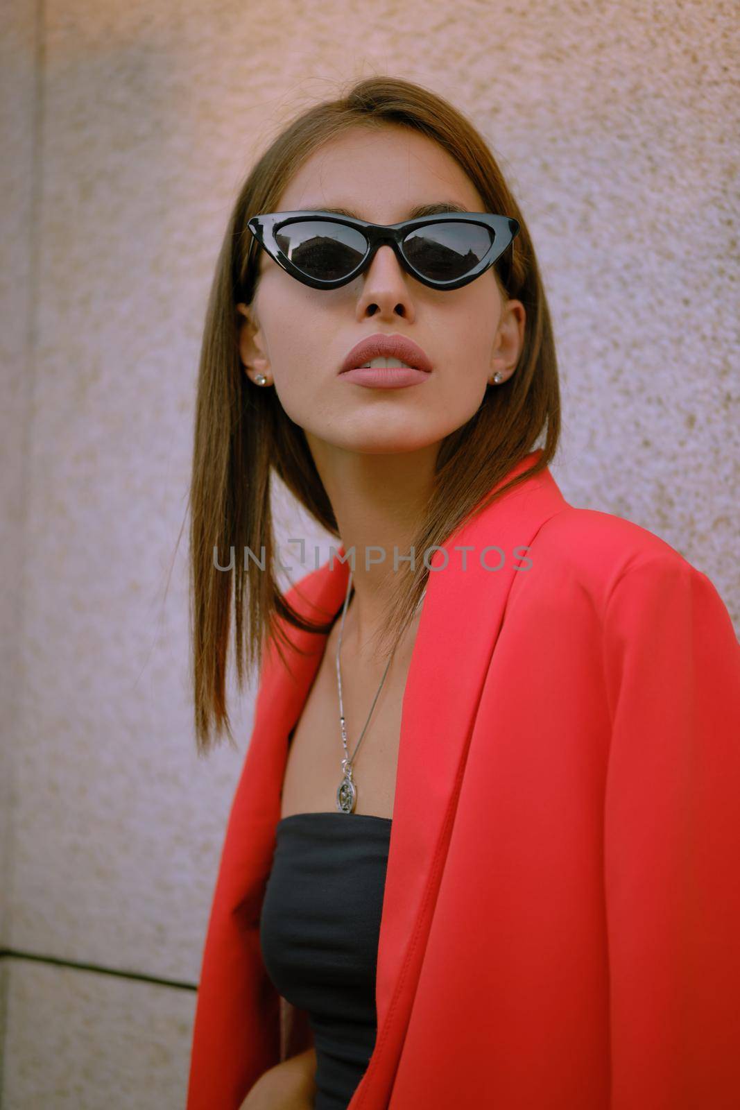 Attractive blonde maiden in a red lady-type pantsuit and black top, sunglasses, with a pendant around her neck is posing near a stone wall. The concept of fashion and style. Close-up shot.