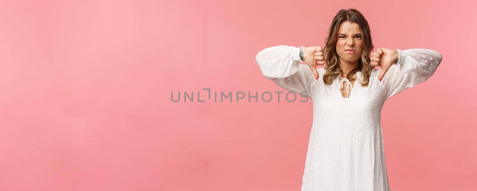 Portrait of picky young blond caucasian woman in white dress over pink background expressing dislike, show thumbs-down and grimacing in aversion, disappointed with bad quality by Benzoix