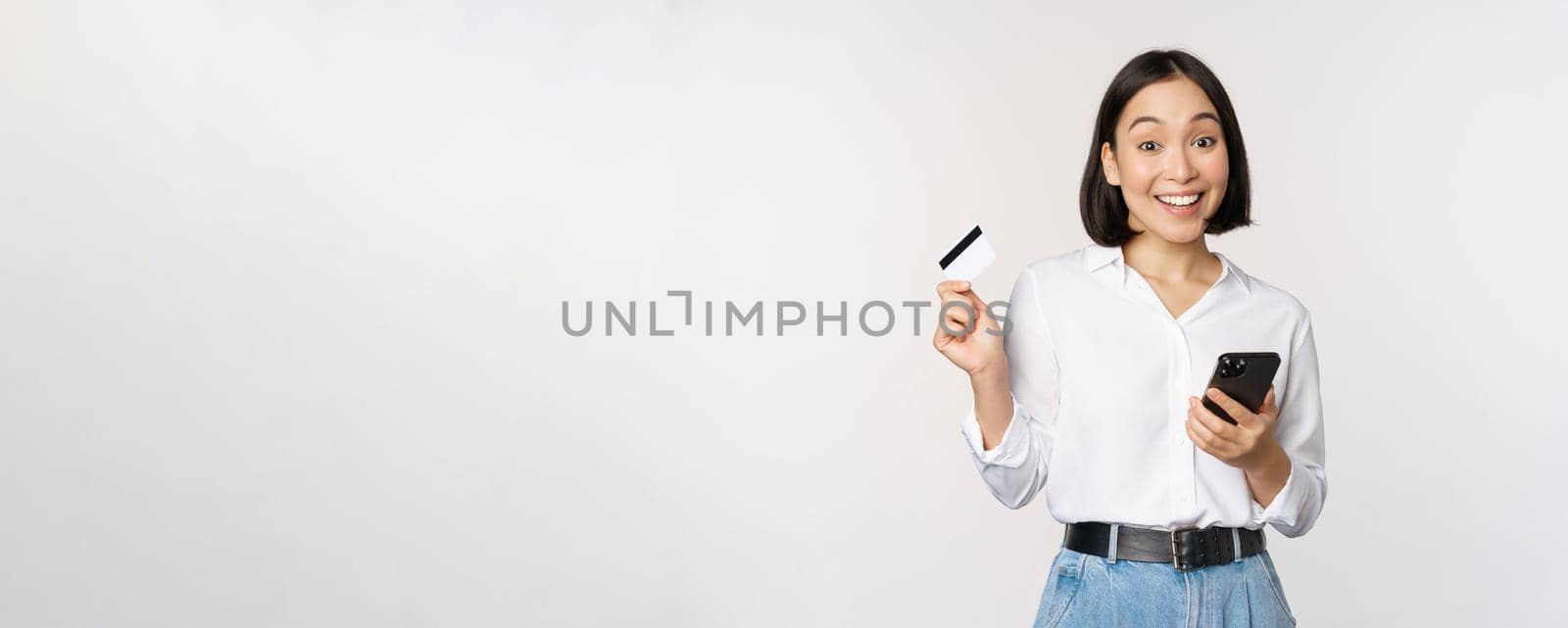 Online shopping concept. Image of young asian modern woman holding credit card and smartphone, buying with smartphone app, paying contactless, standing over white background.