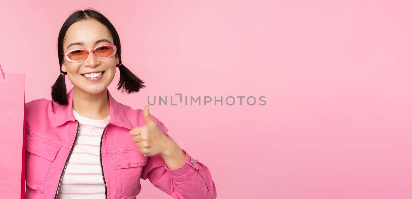 Shopping. Stylish asian girl in sunglasses, showing bag from shop and smiling, recommending sale promo in store, standing over pink background.