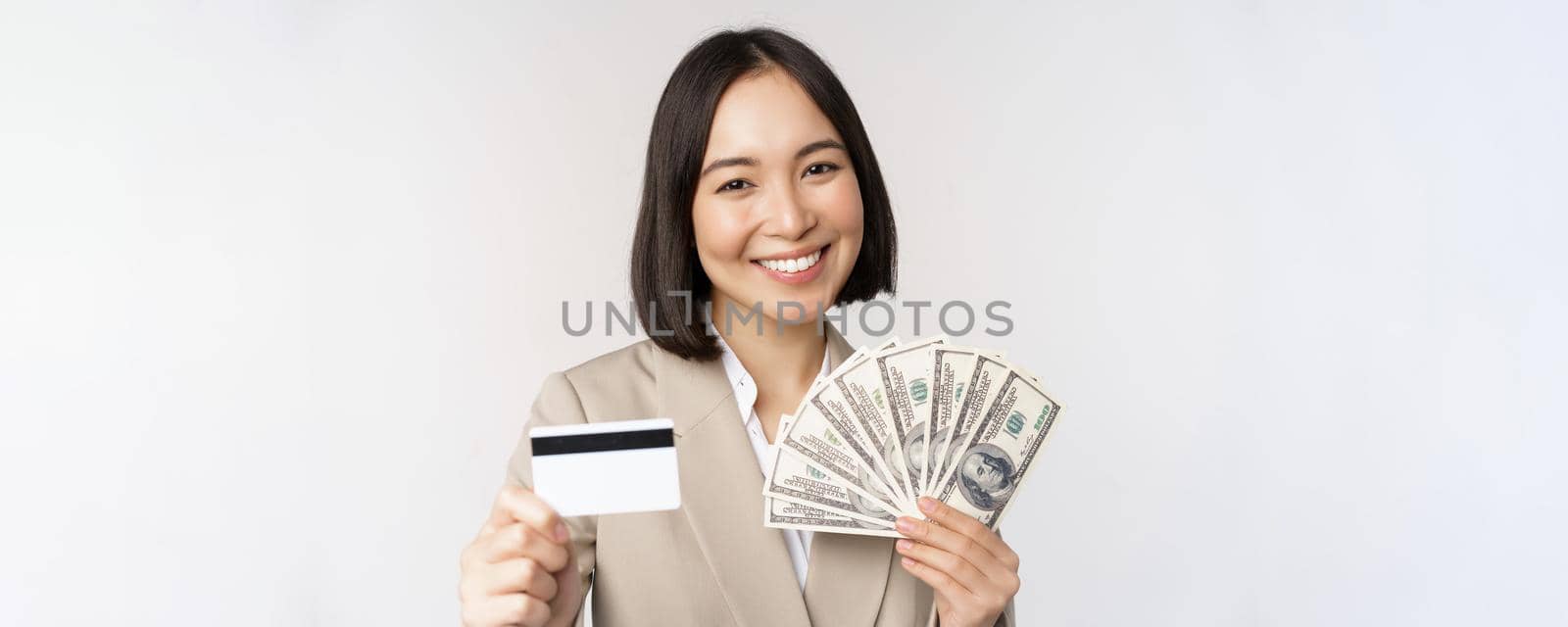 Close up of asian businesswoman, office lady showing credit card and money dollars, standing in suit over white background.