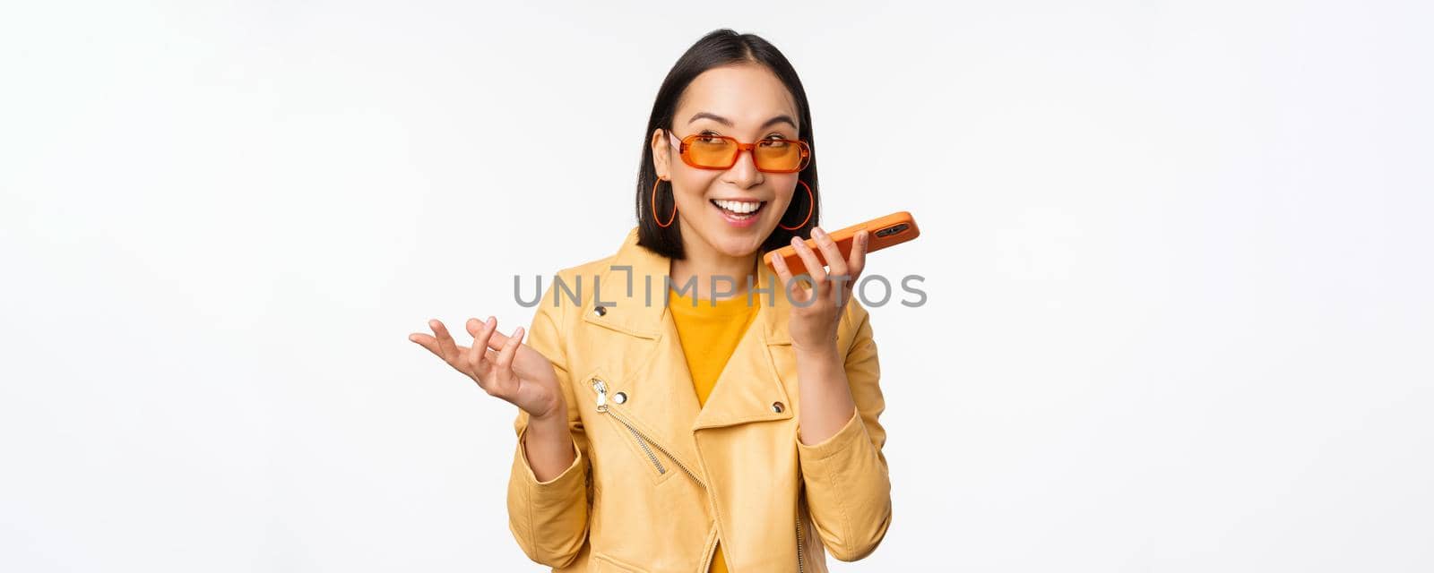 Image of happy asian girl talking on speakerphone, recording, translating her voice with mobile phone app, talking in smartphone dynamic, standing over white background.