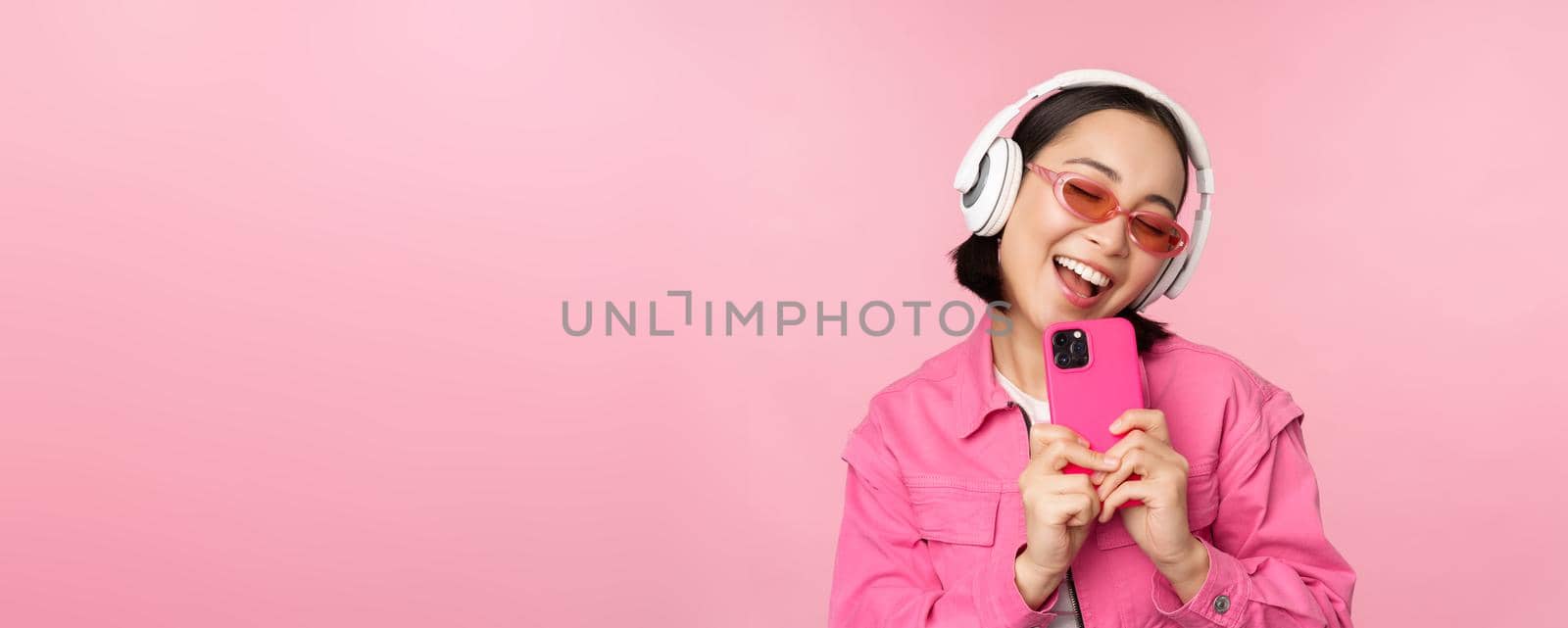 Stylish asian girl in headphones, listening music and taking photos on mobile phone, using smartphone, standing over pink background.