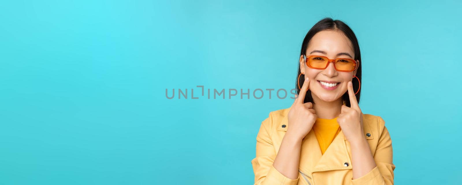 Close up portrait of asian young woman in sunglasses, smiling and looking romantic, standing happy over blue background. Copy space